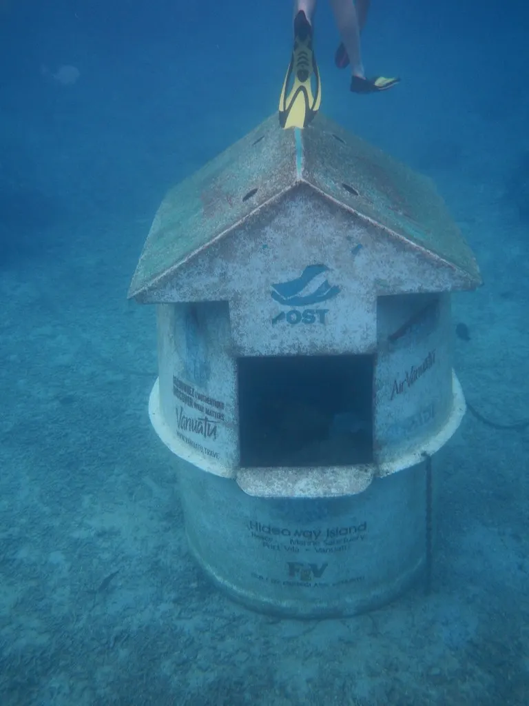 Une petite construction en forme de maison posée au fond sableux, le mot POST et un guichet carré sur sa façade, dans une eau bleue, les palmes jaunes d’un nageur en haut de l’image